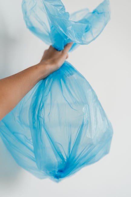 A person's hand holding a large, transparent blue plastic rubbish bag filled with waste, with the bag's opening gathered and pinched at the top. The background is plain and white, emphasizing the focus on the bag and the hand. This image depicts waste disposal which is an essential part of home relocation and moving preparations, often managed by professional removals services like Man with Van Purley. The bag appears to contain household waste, ready for disposal, aligning with waste management rules for residents in Purley. The lighting is even, highlighting the crinkled texture of the plastic, and the scene is set indoors, indicating the process of sorting and disposing of waste materials in preparation for or following a house move.