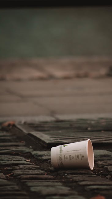 A disposable paper cup with printed text lies on a brick step just outside a doorway, partially tipped over. The surrounding area includes a weathered wooden doorstep, a metal gateway grate, and a paved outdoor surface. The scene is illuminated by natural light, with a background that appears to be a garden or yard area. This image highlights waste disposal concerns relevant to house removals and the importance of proper waste management in the context of property clearance, as part of the services provided by Man with Van Purley for home relocation and furniture transport. The setting illustrates typical environmental considerations involved in moving processes, emphasizing the need for responsible waste disposal in compliance with local regulations.