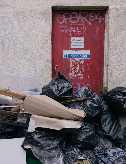 An outdoor scene showing a red metal door with graffiti, signs including one that says 'No Money' and 'Keep Clear,' set within a plain, textured concrete wall. In front of the door, there is a collection of garbage bags, most of which are black plastic, some partially torn or crumpled. Among the bags, there are discarded cardboard boxes, some flattened and others still in shape, along with a plastic container and packaging materials. The area appears cluttered and untidy, with the garbage situated on a paved or concrete surface. The image captures the process of waste collection or disposal outside a building, relevant for professionals involved in house removals or relocation services, especially when considering the logistics of clearing or preparing a property for a move. Man with Van Purley occasionally handles such clearance tasks as part of comprehensive home relocation and furniture transport services, ensuring a clean and ready property for occupancy or further moving phases.