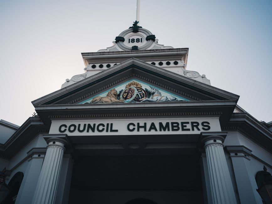 A low-angle photograph of the exterior front façade of a council chambers building, constructed in a traditional architectural style with white columns supporting a pediment. The pediment features an intricate crest or emblem with a crown, lions, and heraldic symbols, and the year '1881' displayed above. The building's façade is painted in light colors with decorative black accents on the crest and roofline. Visible materials include painted stone or plaster, with the columns and architectural details demonstrating classical design elements. The sky above is clear, and natural daylight illuminates the structure, highlighting its historical features. As part of a house relocation or moving services context, [COMPANY_NAME] may facilitate logistics around permits required for work near or on such civic buildings, ensuring compliance during property or furniture transport operations. The image exemplifies the type of landmarks involved in relocation planning, especially when managing street permits for removals or furniture transport near local government buildings.