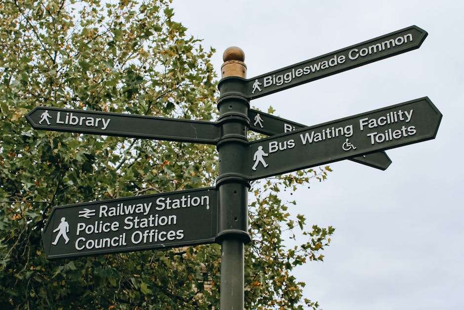 A black multi-directional street signpost situated outdoors against a backdrop of leafy trees with green and brown foliage. The signpost features six signs pointing in various directions; from top to bottom, signs indicate Biggleswade Common, Library, Bus Waiting Facility and Toilets, Railway Station, Police Station, and Council Offices. The signs have white text and icons, with some including pedestrian symbols and accessibility icons. The sky overhead is overcast, providing diffuse, natural lighting. The scene illustrates a typical location for navigating services and amenities within a residential or town area, relevant to house removals and local relocation processes, highlighting the importance of navigation and signage in community mobility and logistics managed by [COMPANY_NAME] for home moving and furniture transport services.