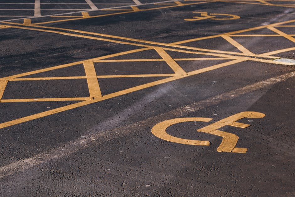 A close-up view of an empty outdoor parking area with a designated space for disabled parking, marked by a yellow wheelchair symbol painted on the dark asphalt surface. Surrounding the disabled parking space are yellow diagonal and horizontal line markings indicating restrictions or specific parking zones. The surface shows signs of wear and slight texture variation. This outdoor scene captures part of the parking area near a residential or commercial property, suitable for the loading process involved in home relocation or furniture transport, as provided by Man with Van Purley for house removals and moving services near Purley Station.