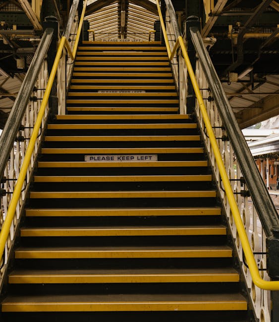 A straight flight of metal stairs with yellow safety railings on both sides, leading up to an indoor platform or loading area within a building. The stairs have black tread surfaces with yellow edges, and the top of the stairs features a paneled ceiling with industrial lighting. A small sign on one of the steps reads 'PLEASE KEEP LEFT.' Surrounding the stairs, visible structural elements include pipes and metal beams, indicating an industrial or warehouse setting, which is relevant to house removals and furniture transport services provided by Man with Van Purley. The lighting is bright, ensuring clear visibility of the entire staircase and its surroundings, emphasizing the logistics involved in moving and packing items within such environments.