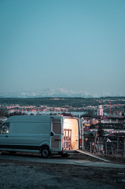 A light grey moving van parked on a slightly inclined paved area with its rear doors open, revealing furniture wrapped in protective materials and cardboard boxes within. The van is positioned to the