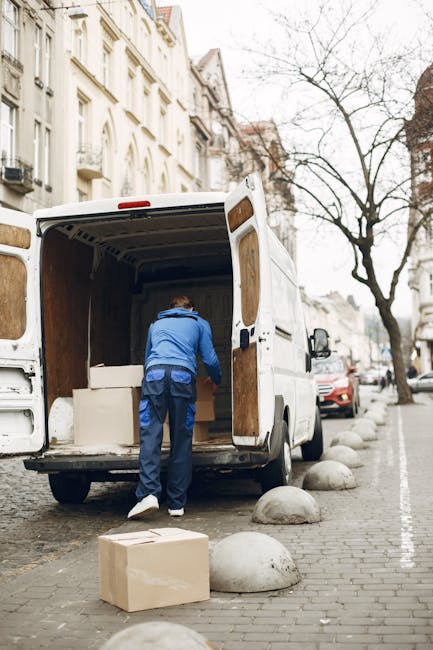 A light grey moving van parked on a slightly inclined paved area with its rear doors open, revealing furniture wrapped in protective materials and cardboard boxes within. The van is positioned to the