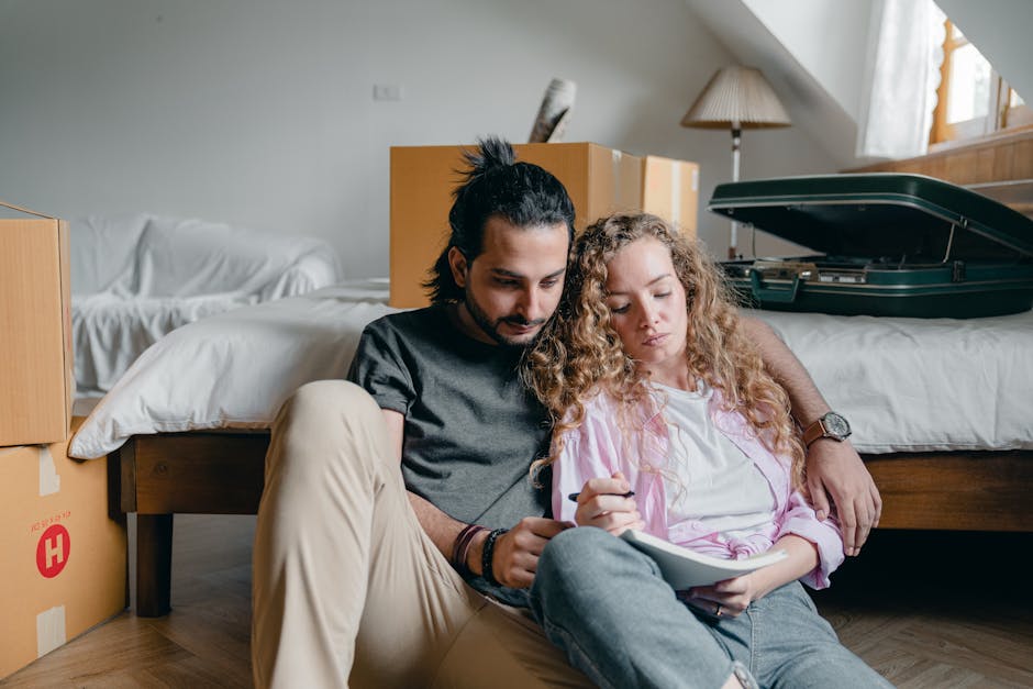 A man with long dark hair tied back and a beard, wearing a black T-shirt and beige trousers, sits on the wooden floor leaning against a bed frame in a bedroom. Beside him, a woman with curly blonde ha