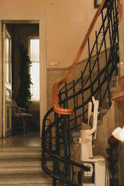 The image shows a staircase inside a home, featuring a decorative black metal balustrade with intricate scrollwork and a polished wooden handrail. The stairs are made of stone or marble, ascending from the ground floor to an upper level. Next to the staircase is a white stairlift with a beige seat and safety harness, positioned on the right side, indicating accessibility considerations during a home relocation or furniture transport. Visible in the background is a doorway leading into a room lit by natural light from a large window, with a radiator beneath it and a potted plant visible on the windowsill. Near the base of the stairs, on the landing, there are cardboard boxes and packing materials, such as bubble wrap and plastic bags, suggesting packing or preparations for moving. The environment appears clean and organized, with neutral-toned walls and minimal clutter, typical of a house being prepared for or undergoing a household move, exemplifying professional removals services by Man with Van Purley.