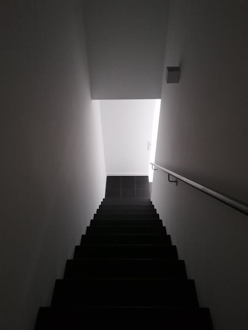 A photograph taken from the top of a staircase in a residential property showing dark carpeted steps descending toward a small landing with a tiled floor. The staircase is enclosed by plain white walls on both sides, with a metal handrail fixed to the right wall for support during navigation. The top landing is lit by natural light entering through a doorway at the bottom, which is open, revealing a bright space beyond. The image captures the narrow, enclosed nature of the staircase, which is part of a house undergoing a home relocation or furniture transport process, as managed by Man with Van Purley, with the focus on the structural details of the stairs and environment in preparation for moving logistics.