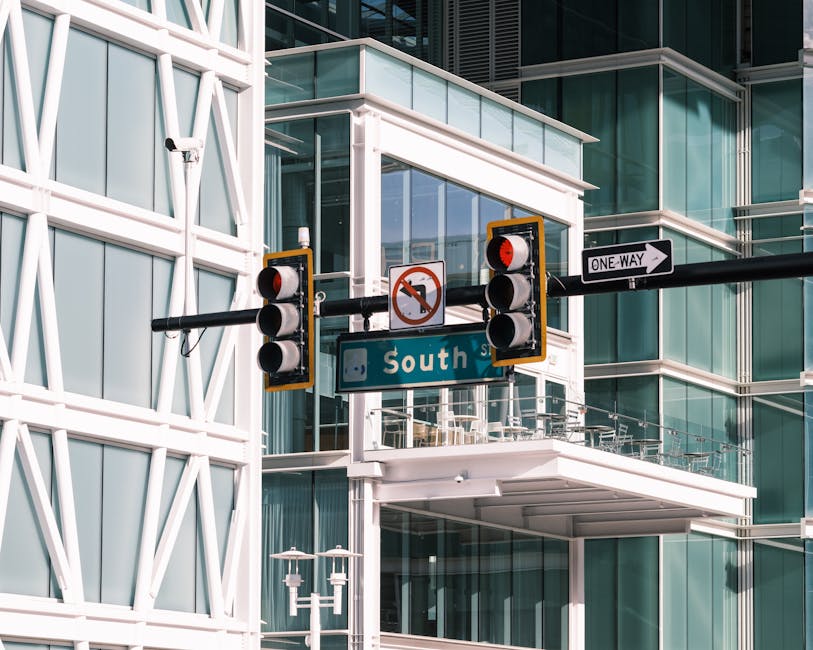 Photograph of a modern urban street scene showing a large traffic light with five circular signals mounted on a black horizontal bar. The signals are red, indicating stop, and one of the lights features a no-turn prohibition sign. Positioned below and slightly behind the traffic light is a street sign labeled 'South St,' attached to the same black bar. To the right, a white one-way sign with a black arrow points to the right. The background showcases a contemporary glass and steel building with multiple balconies, some with glass railings, and sunlit windows reflecting the sky. The scene suggests an active city area, possibly near office or residential buildings, with the traffic control equipment regulating local vehicle movement. The image is in a well-lit environment, capturing the urban infrastructure essential for city traffic and pedestrian flow, which aligns with the themes of relocation logistics and moving services offered by Man with Van Purley, especially when planning office moves or large-scale house removals.