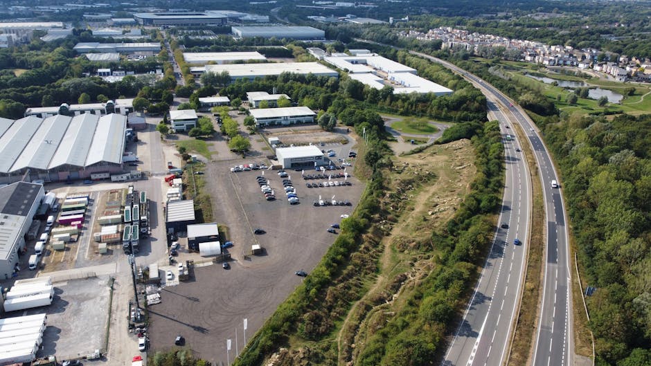An aerial view of an industrial estate adjacent to a dual carriageway, showing multiple large warehouse buildings with white and grey roofs, surrounded by open parking lots filled with cars and trucks. In the foreground, there are several trucks and trailers parked near loading bays, with some equipment such as pallets and storage containers visible. To the right, a green, wooded embankment runs alongside the road, with dense trees and shrubs indicating the boundary of the estate. The overall scene is well-lit with natural daylight, illustrating a busy industrial area that could be involved in logistics and furniture transport, relevant to house removals or moving services. This image exemplifies the logistics planning and infrastructure utilized by companies like Man with Van Purley during packing and loading for a house relocation or business move, as part of comprehensive moving and removal solutions.
