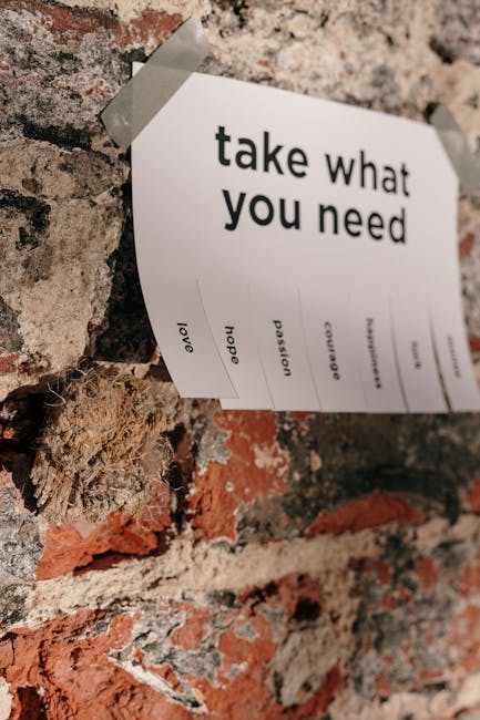 A white paper note taped with clear adhesive tape to a textured brick wall, displaying the message 'take what you need' in bold black letters. Below this message, various words such as 'hope,' 'passion,' and 'love' are printed in smaller font. The bricks are a mix of reddish-brown, cream, and black hues, with some mortar visible. The note is positioned slightly askew, and the wall surface appears rough and weathered, providing a contrasting background for the message. This image relates to themes of emotional support and motivation, suitable for content about house removals and relocation services offered by Man with Van Purley, aligning with home moving and packing processes.