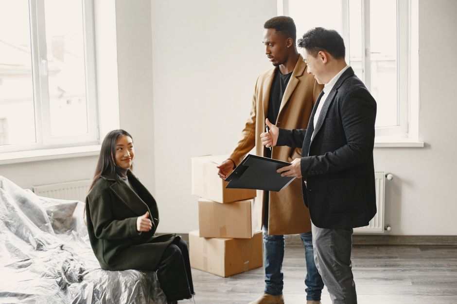 Inside a bright, minimally furnished room with large windows allowing natural light, a woman is seated on a bed covered with a grey satin sheet, looking towards two men who are standing nearby. The men are engaged in a discussion, with one holding a black clipboard and gesturing towards the woman. Behind them, there are several packed cardboard boxes stacked on the floor, ready for transportation. The scene captures the process of home relocation or packing as part of a removal service, with the men possibly representing movers from Man with Van Purley, preparing for a furniture transport or loading process as part of an urgent eviction or house move in Purley. The room's clean appearance and the presence of packing materials highlight a professional and organized approach to house removals.