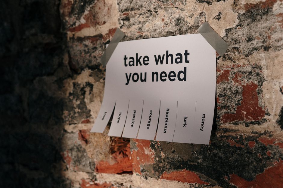A white paper note taped with clear adhesive tape to a textured brick wall, displaying the message 'take what you need' in bold black letters. Below this message, various words such as 'hope,' 'passion,' and 'love' are printed in smaller font. The bricks are a mix of reddish-brown, cream, and black hues, with some mortar visible. The note is positioned slightly askew, and the wall surface appears rough and weathered, providing a contrasting background for the message. This image relates to themes of emotional support and motivation, suitable for content about house removals and relocation services offered by Man with Van Purley, aligning with home moving and packing processes.