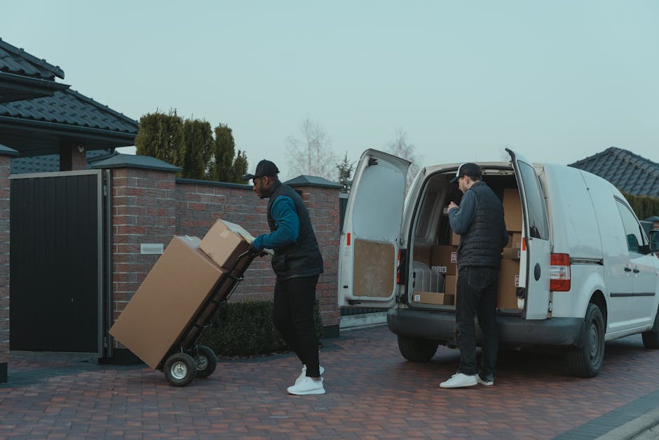 Two men are engaged in furniture transport and packing for a home relocation on a residential driveway. One man is pushing a small black trolley loaded with large cardboard boxes, lifting a box onto the trolley, while wearing a navy jacket, black trousers, white sneakers, and a cap. The other man is standing at the open rear doors of a white cargo van parked on the brick driveway, organizing or loading additional cardboard boxes inside. The van's interior is visible, containing more boxes and packing materials. The scene is set outside in a quiet neighborhood with brick and tiled houses, a black gate, and a fence, under a cloudy sky. The environment appears well-lit with natural daylight, and the overall activity reflects movement related to packing and furniture transport during a house removal, with Man with Van Purley providing these moving services.