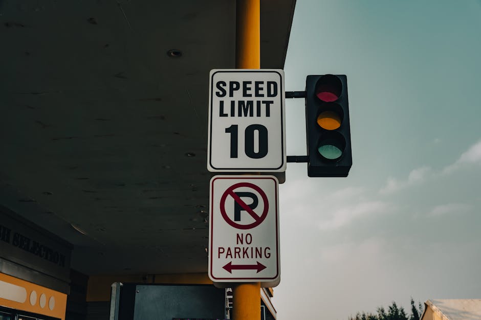 A vertical pole mounted on a pavement in an urban setting displays two traffic signs: the upper sign indicates a speed limit of 10 miles per hour with a white background and black text, attached to a black traffic light showing a red signal; the lower sign is a no parking notice with a white background, a red circle with a slash through a letter 'P', and red text stating 'NO PARKING' with arrows pointing left and right. The background features part of a building with a yellow and black awning, a cloudy sky, and some trees in the distance. This scene captures elements related to street regulation and parking restrictions, relevant for house removals and moving services such as loading and parking logistics, as depicted in the page about 'Van Loading & Parking Rules on Purley High Street, PURLEY' at manwithvanpurley.co.uk.